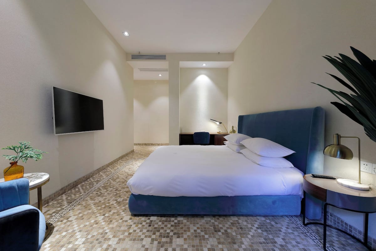 A hotel room with a large bed, a flat-screen TV mounted on the wall, a small table with a lamp, and a potted plant.