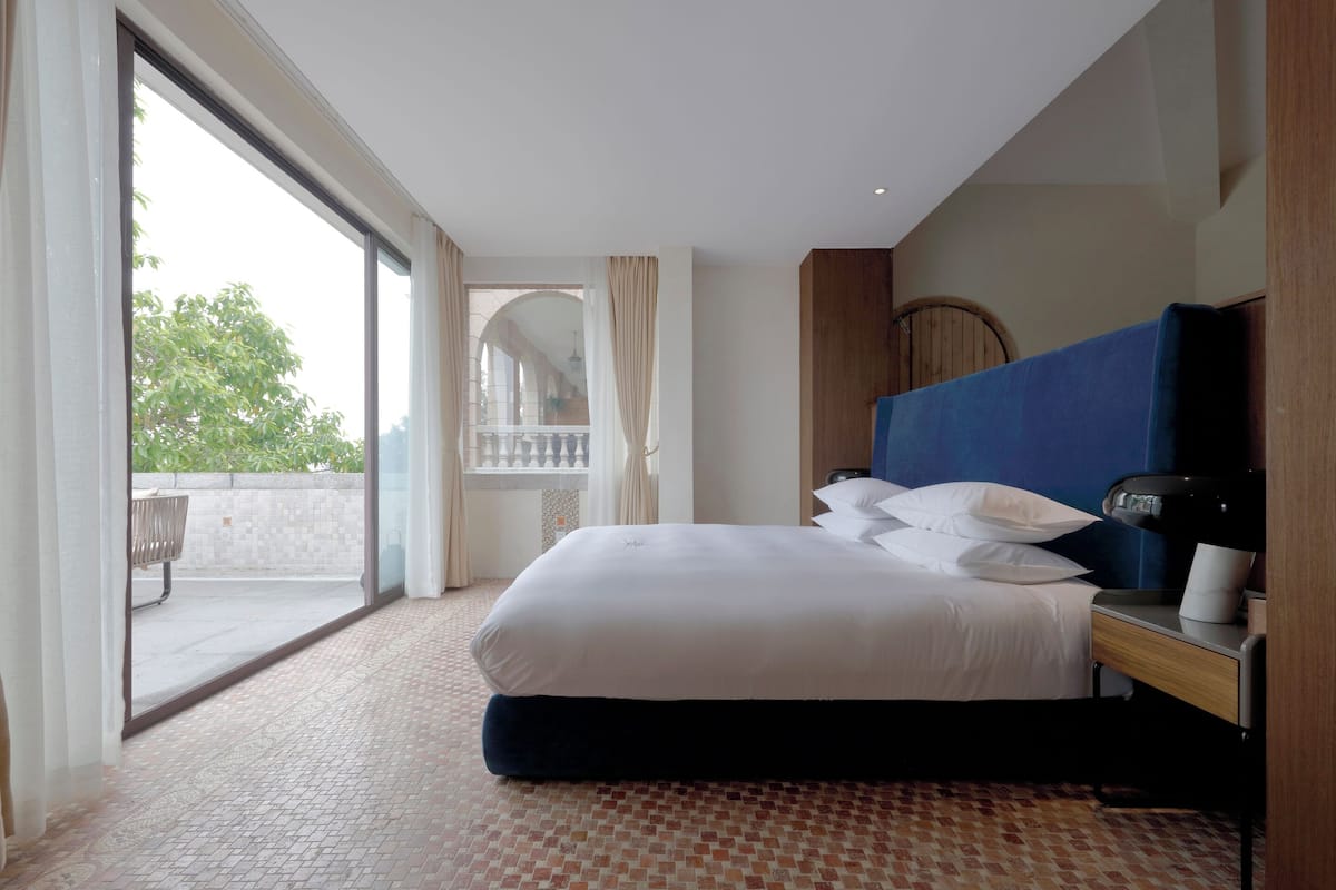 A hotel room with a large bed, a desk with a lamp, and a view of the outdoors through a sliding door.