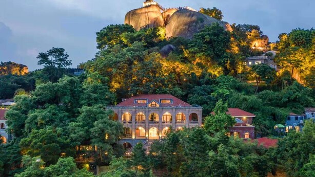 A large building with multiple arches and a red roof, surrounded by dense greenery and a prominent rock formation in the background. - Living Sense Linghai Hotel (Xiamen)