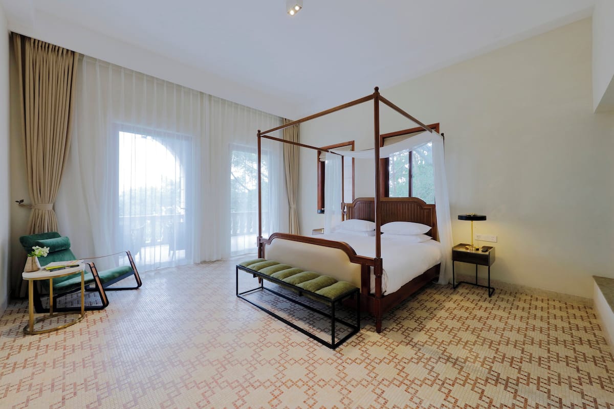 A four-poster bed with a bench, a green armchair, a small table, and a nightstand in a hotel room with patterned carpet and large windows.