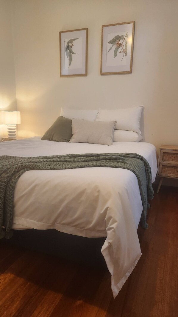 Una cama bien hecha con ropa blanca y un plaid verde de punto. Encima de la cama, en la pared, hay dos cuadros con motivos botánicos.