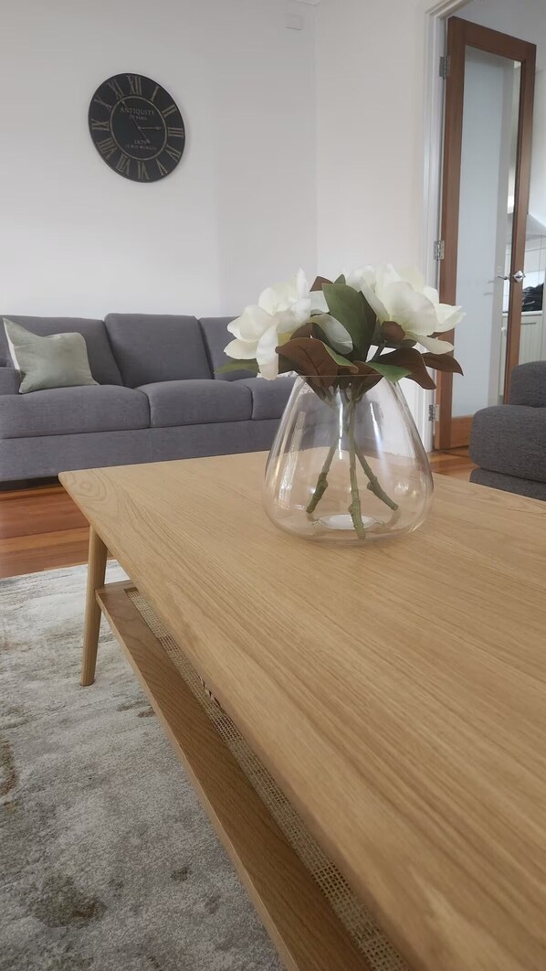A living room with a grey sofa, a wooden coffee table with a vase of white flowers, and a wall clock.