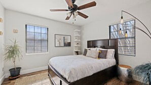 Un dormitorio con cama, ventilador de techo, planta en maceta, mesita de noche y ventana con persianas.