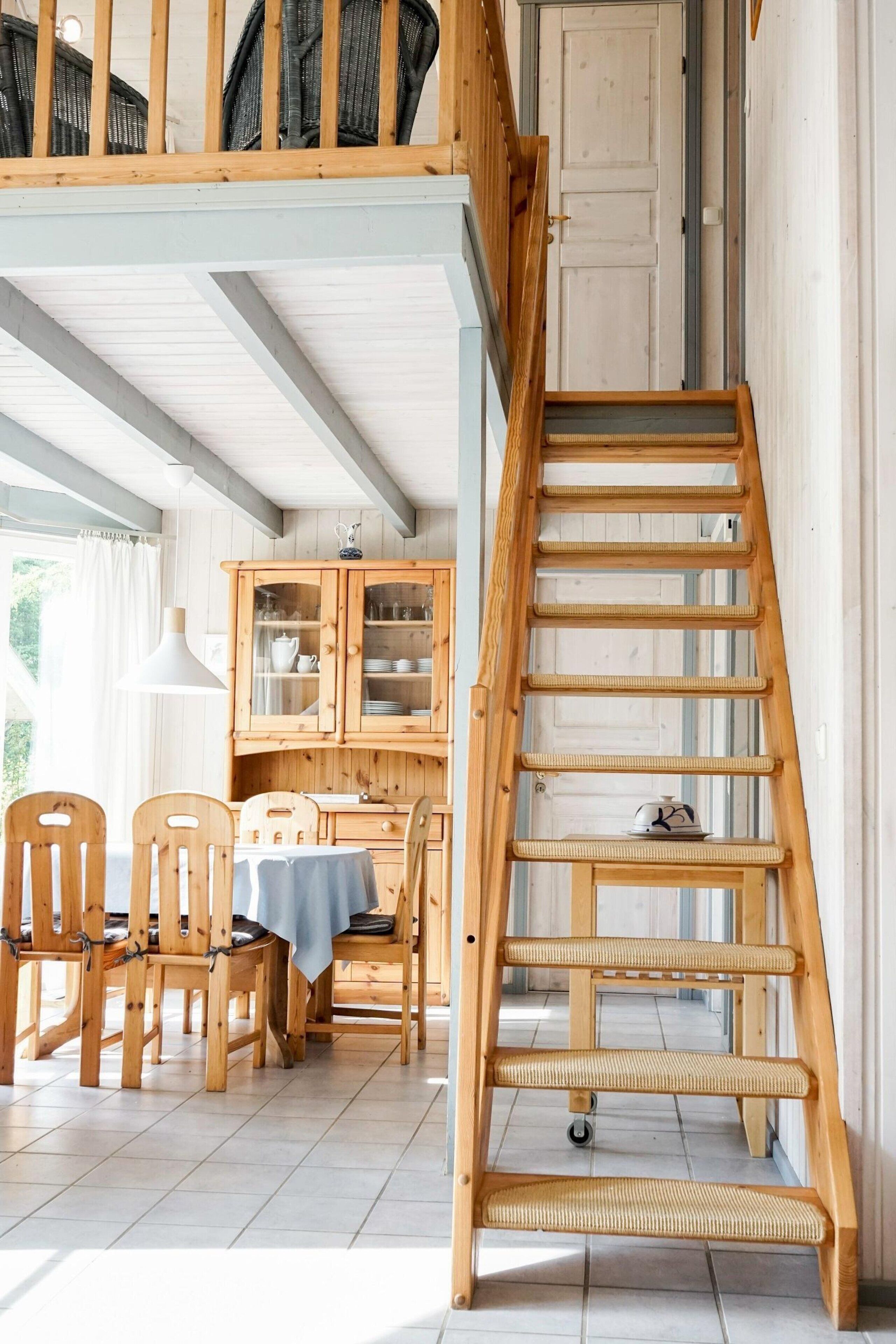 Un escalier en bois avec un petit panier à sa base, un coin repas avec des chaises et une table en bois, et une cuisine avec des placards en bois.