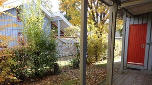 A covered outdoor area with a wooden deck, a red door, and a building with a sloped roof in the background. - Ferienhaus im Skandinavischen Stil (Mirow)