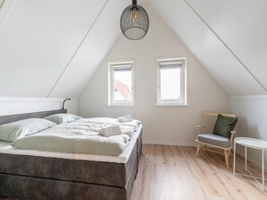 Ein Schlafzimmer mit einem Bett, zwei Fenstern, einem Sessel, einem kleinen Tisch und einer Pendelleuchte.
