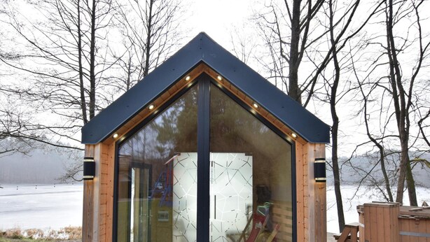 A glass-enclosed wooden cabin with a sloped roof, a wooden deck, and a view of a lake.