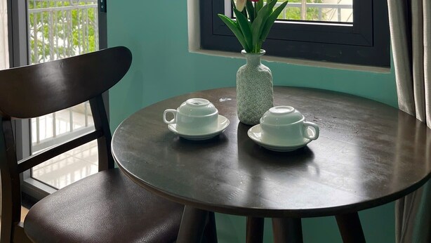 A room with a round table, two white teacups, a vase with tulips, a chair, and a window with a view of houses. - Bunk Beach Hostel (Da Nang)