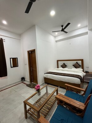 A modern hotel room with a large bed, a wooden coffee table, a blue sofa, a ceiling fan, and a small table with a vase of flowers. - SRH HOTEL (Greater Noida)
