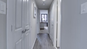 Un pasillo estrecho con puertas blancas a ambos lados, que conduce a una zona de cocina con ventana y una lámpara colgante.