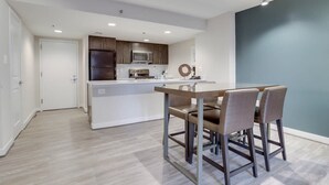 Una cocina moderna con armarios oscuros, un refrigerador de acero inoxidable y un microondas. Hay un comedor con una mesa de vidrio y dos sillas.