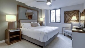 Una habitación de hotel moderna con una cama grande, un escritorio, una silla, un ventilador de techo, un ventanal y un panel decorativo en la pared.