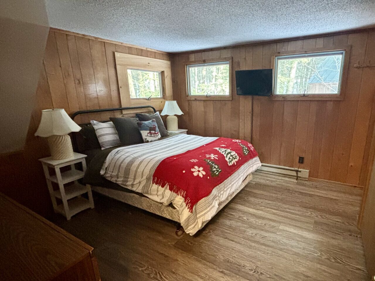A bedroom with a wooden floor, a bed with a red blanket, two bedside tables with lamps, and two windows.
