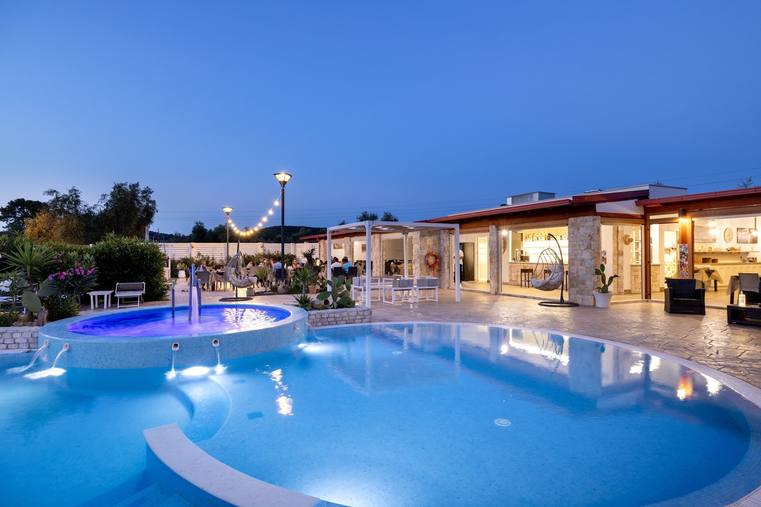 A swimming pool with a water fountain, surrounded by outdoor seating and a restaurant.