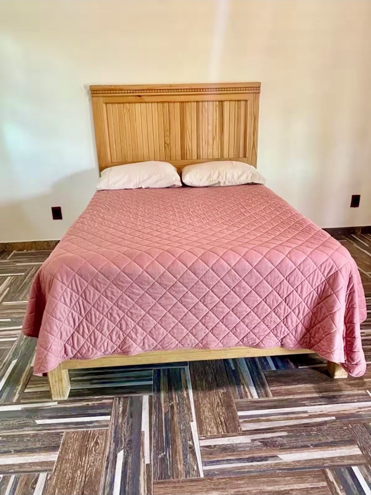 Un lit avec une tête de lit en bois, une couvre-lit rose matelassée et deux oreillers blancs. Le sol est recouvert d’un parquet à motifs en chevrons, dans différentes nuances de bois.