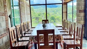 A long wooden dining table with chairs, set against a brick wall with large windows offering a view of a green landscape.
