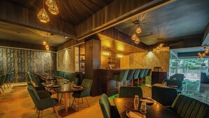 Un restaurant avec des chaises et des tables recouvertes d’un tissu vert, des suspensions lumineuses et une vue sur l’extérieur grâce à de grandes fenêtres.
