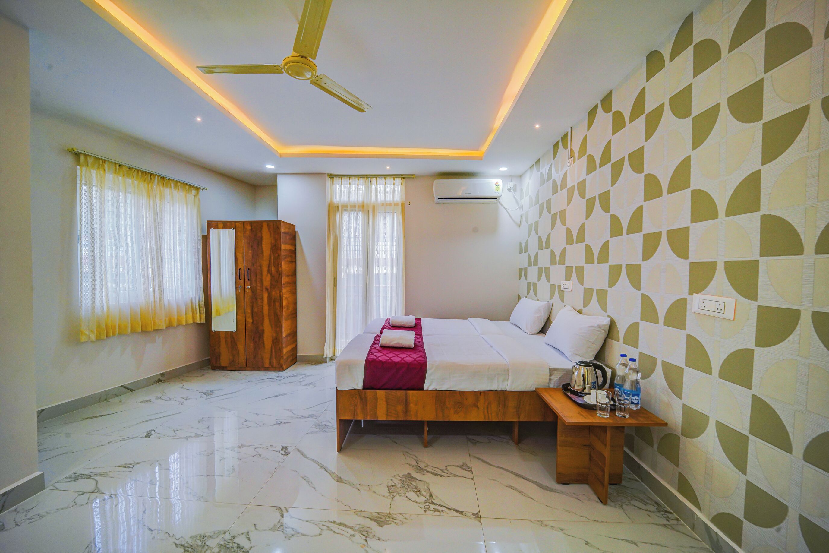 A hotel room with a bed, a wooden wardrobe, a bedside table with a lamp, and a ceiling fan.