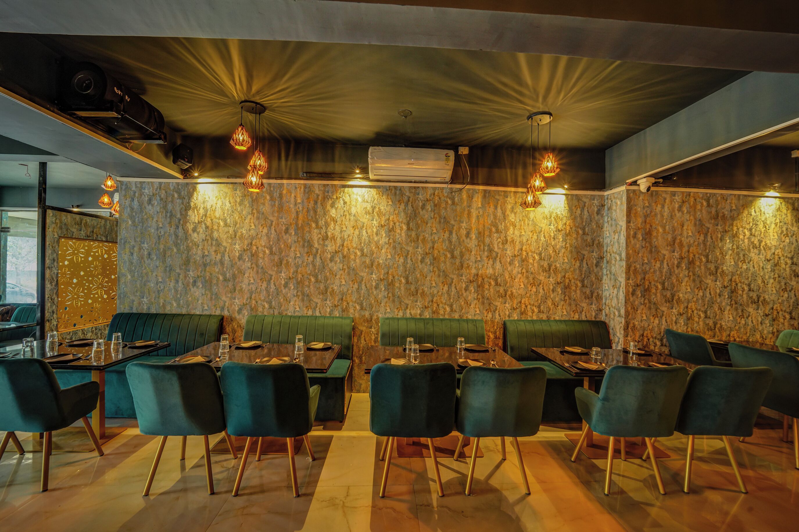 Un restaurant avec des banquettes et des tables recouvertes d’un tissu vert, un éclairage chaleureux et un mur à la texture intéressante.