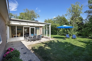 Une terrasse couverte avec une table et des chaises, un parasol bleu et deux fauteuils de jardin, le tout situé dans un jardin.