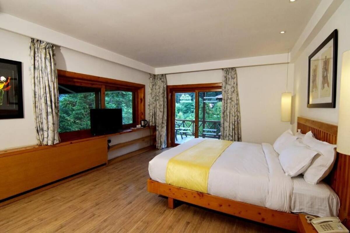 A bedroom with a wooden bed, a TV on a wooden bench, and large windows with curtains.