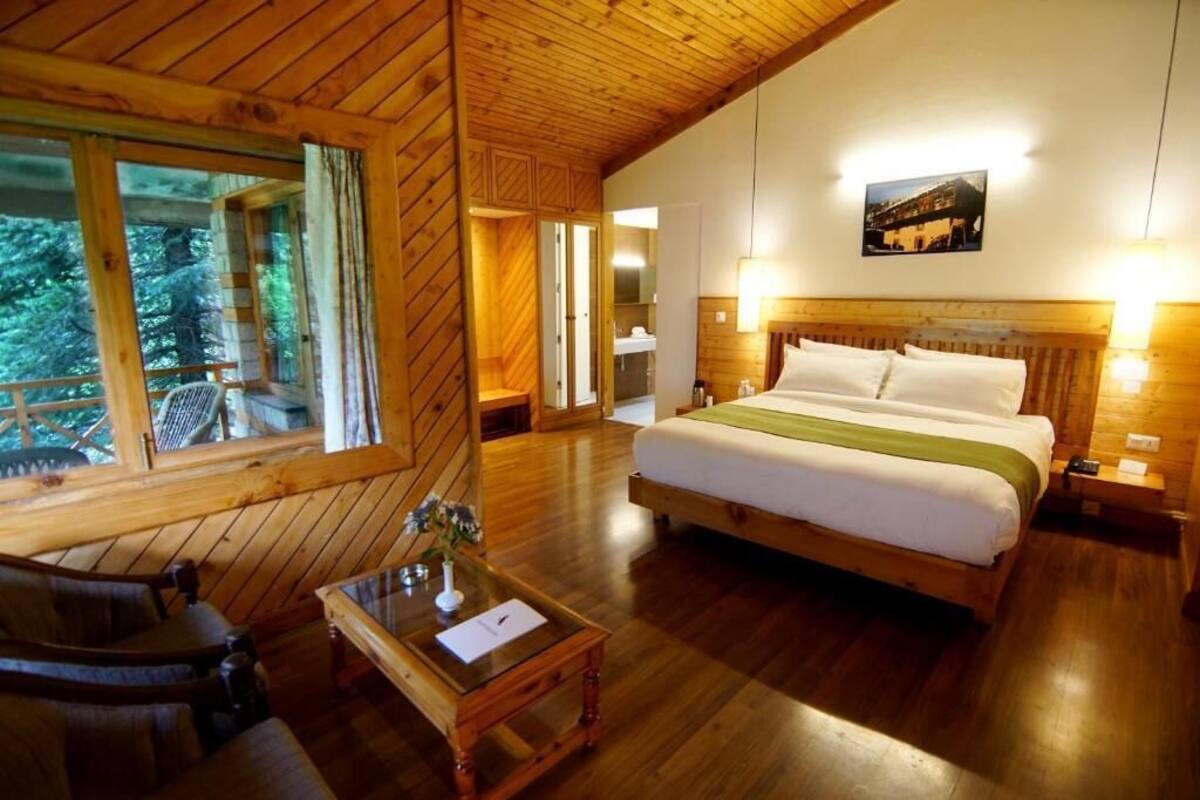 A wooden cabin room with a large bed, a wooden desk, and a window overlooking a forest.
