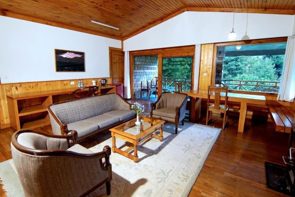 A cozy living room with a sofa, armchairs, and a coffee table. There is a large window with a view of greenery outside.