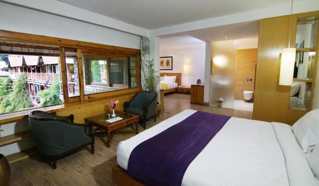 A hotel room with a large bed, a desk, a chair, a small table, and a window with a view of a wooden deck and greenery.