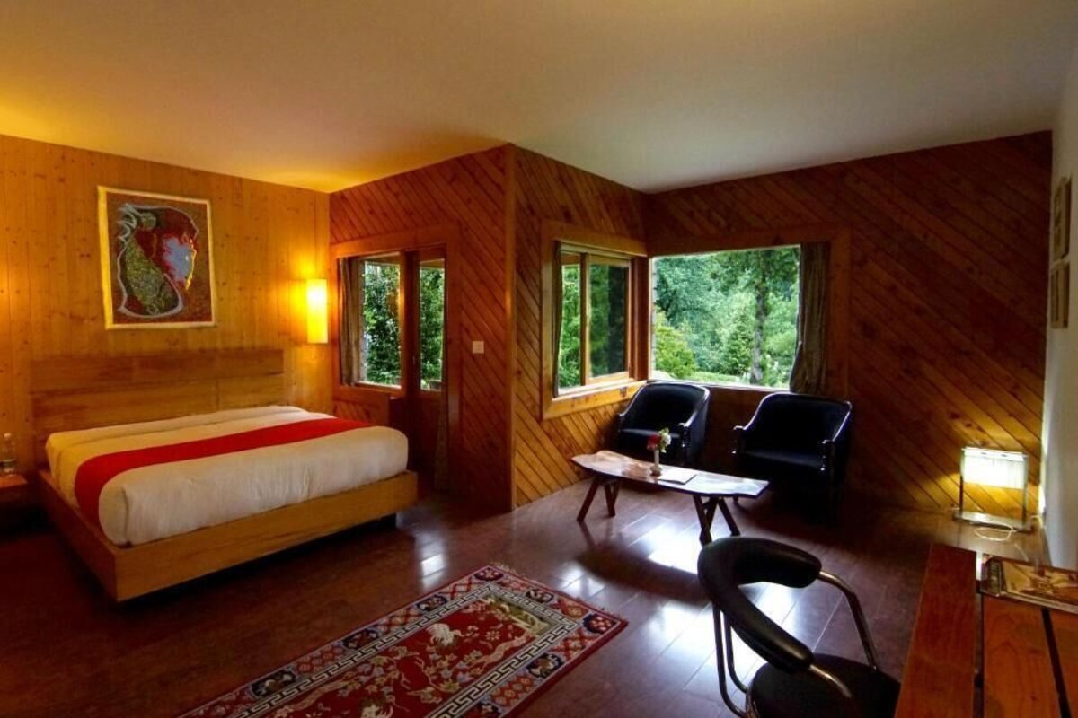 A wooden-themed room with a bed, two chairs, a small table, and large windows offering a view of greenery.