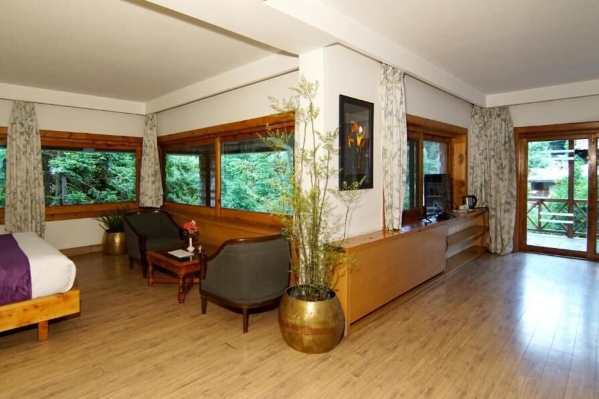 A room with a bed, a desk, a chair, and a large window overlooking a forest.