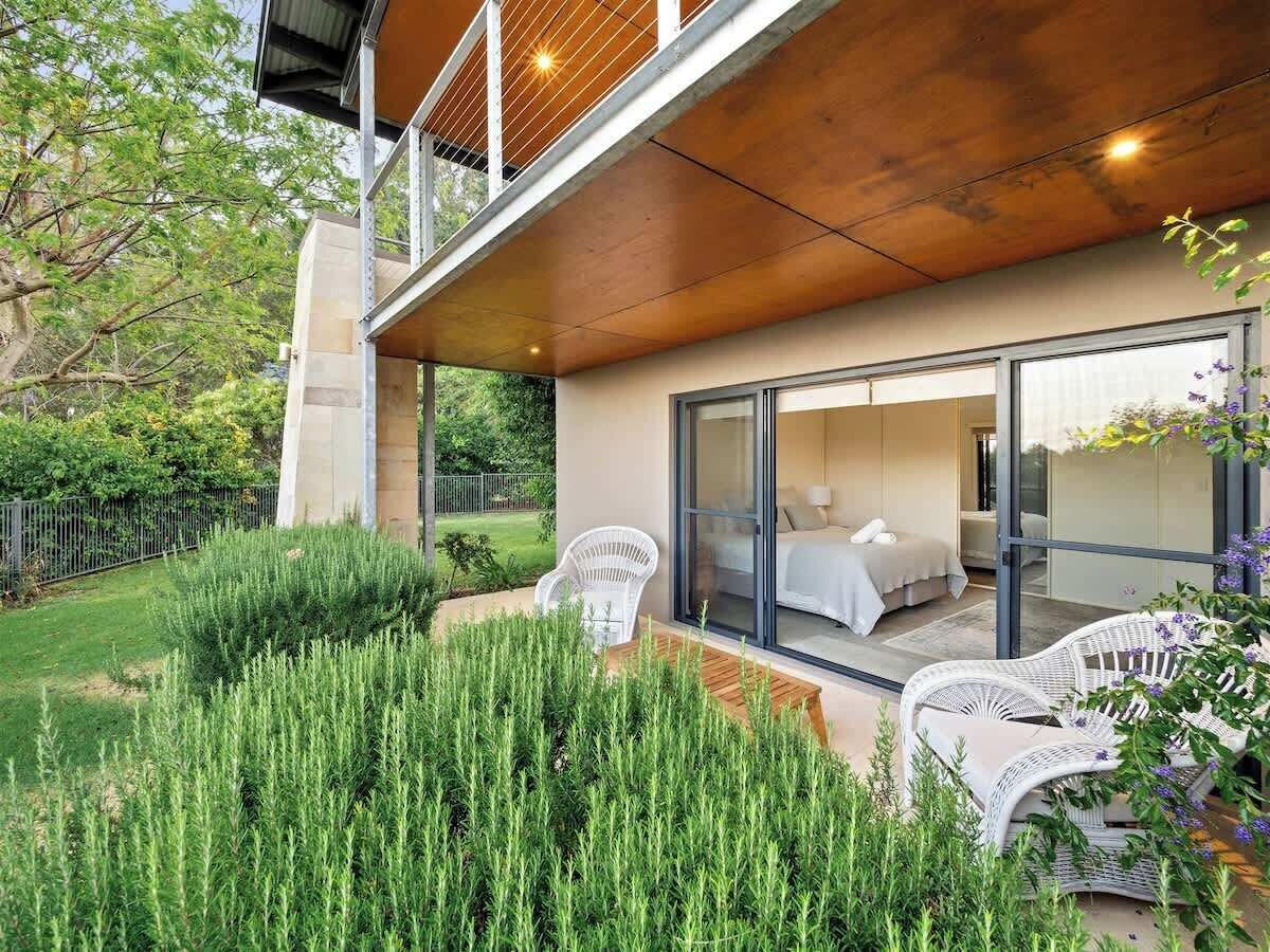A modern house with a large glass door leading to a bedroom, a balcony with outdoor furniture, and a well-maintained garden.
