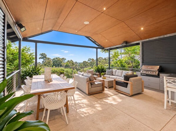 An outdoor patio with a wooden ceiling, a dining table with chairs, a sofa, and a grill.