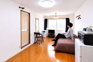 A compact living space with a bed, a television, a small table with a chair, a window with curtains, and a kitchenette area. - Higashi-Sapporo Residence minpaku (Sapporo)