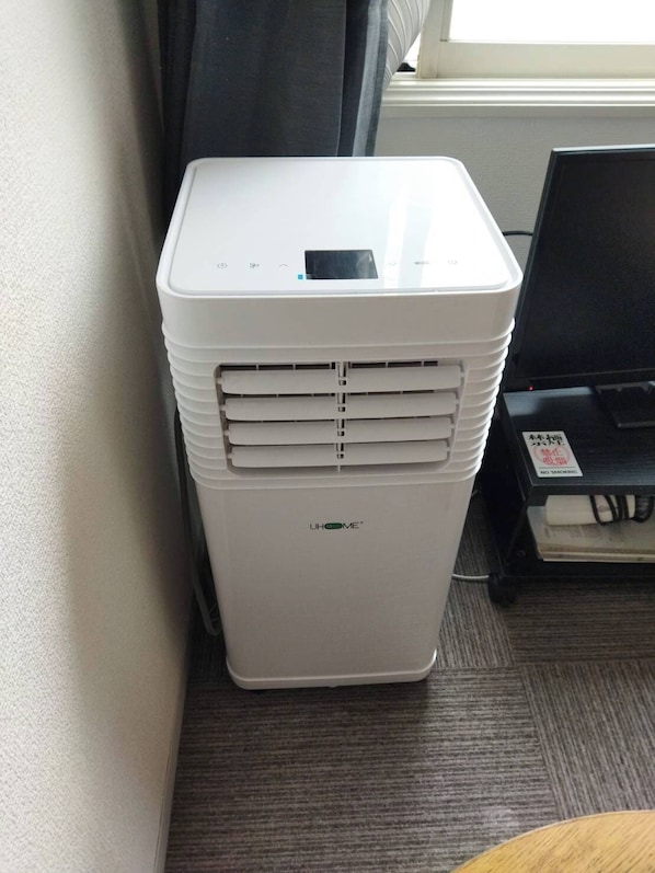 A white portable air conditioner with a control panel, placed on a carpeted floor next to a window with curtains. - Higashi-Sapporo Residence minpaku (Sapporo)