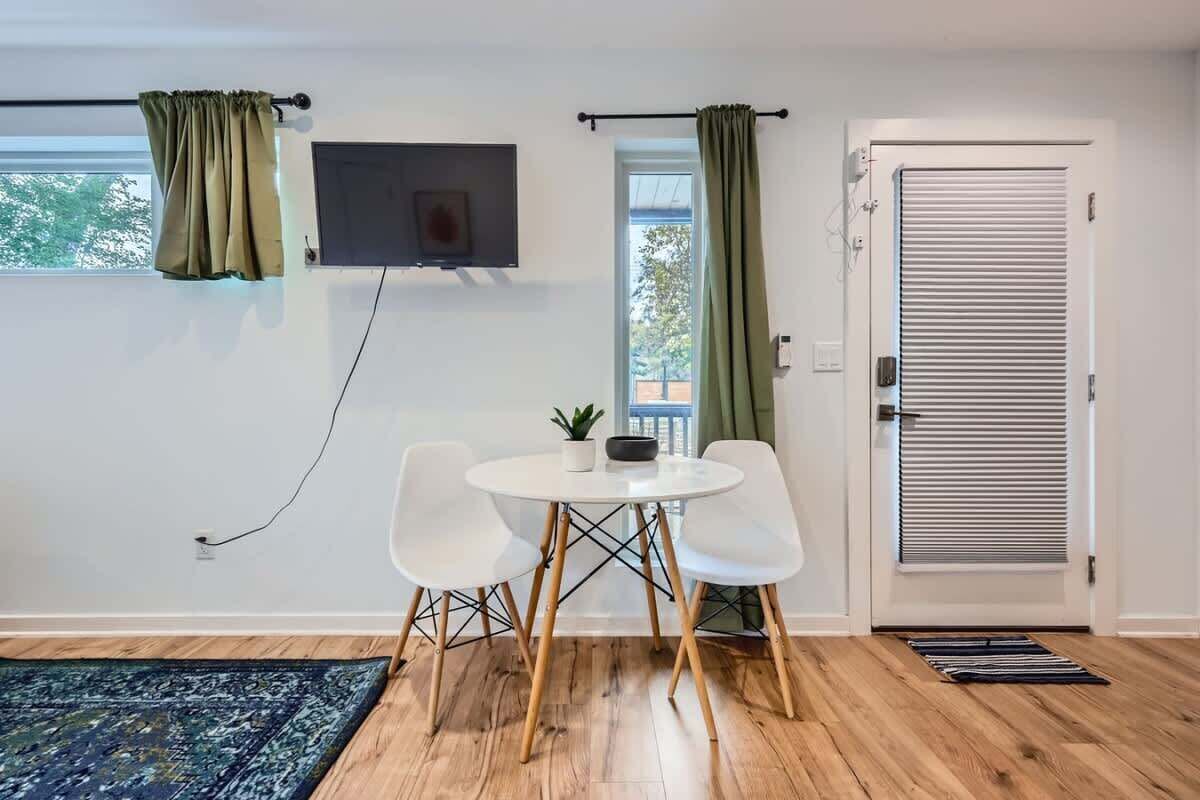 A room with a white table and chairs, a flat-screen TV mounted on the wall, a sliding door with a window, and a patterned rug on the floor.