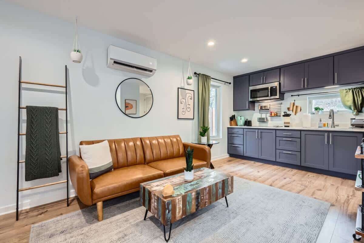 Een moderne woonkamer met een bruine leren bank, een salontafel met een rustieke uitstraling, en een keukenhoek met donkere kastjes en een magnetron.