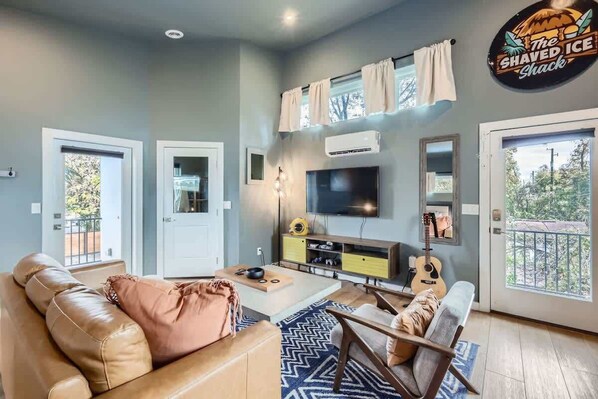 A living room with a beige sofa, a coffee table, a TV, and a guitar. There are two doors, one leading outside and another to a balcony. - Holly Studio walkable to Downtown, bars, SXSW (Austin)