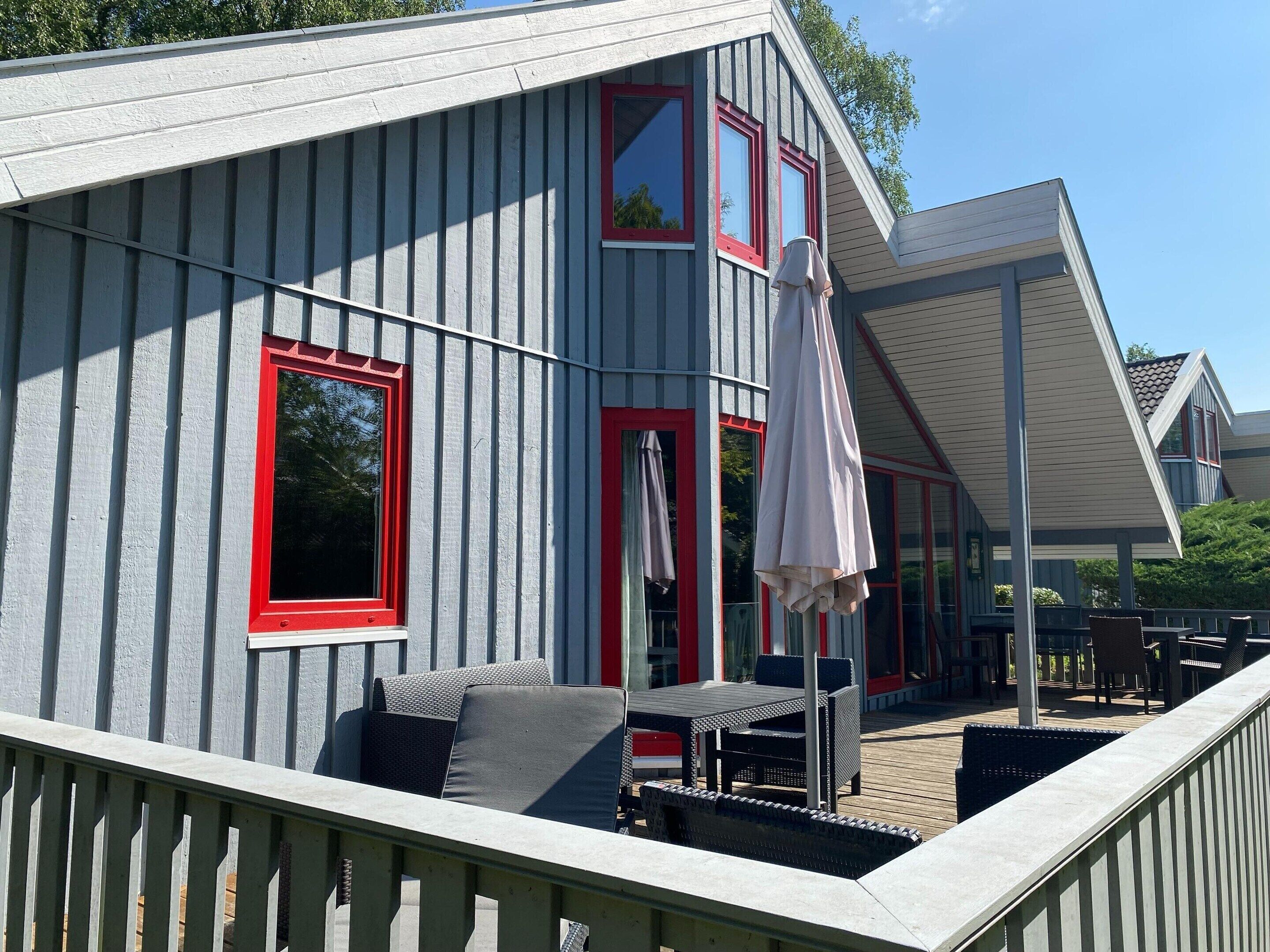 A covered outdoor seating area with a table and chairs, a pergola, and a view of a building with red-framed windows.