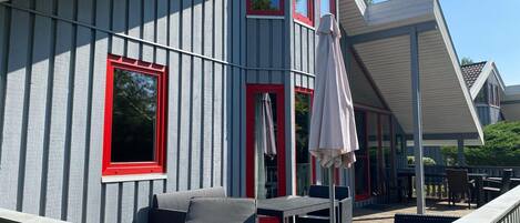 A covered outdoor seating area with a table and chairs, a pergola, and a view of a building with red-framed windows.