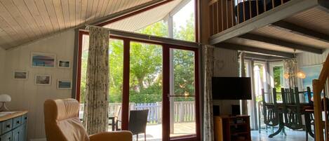 Un vaste salon doté d’un haut plafond, une porte-fenêtre coulissante donnant sur un balcon, un coin repas avec une table et des chaises, et un escalier en bois.