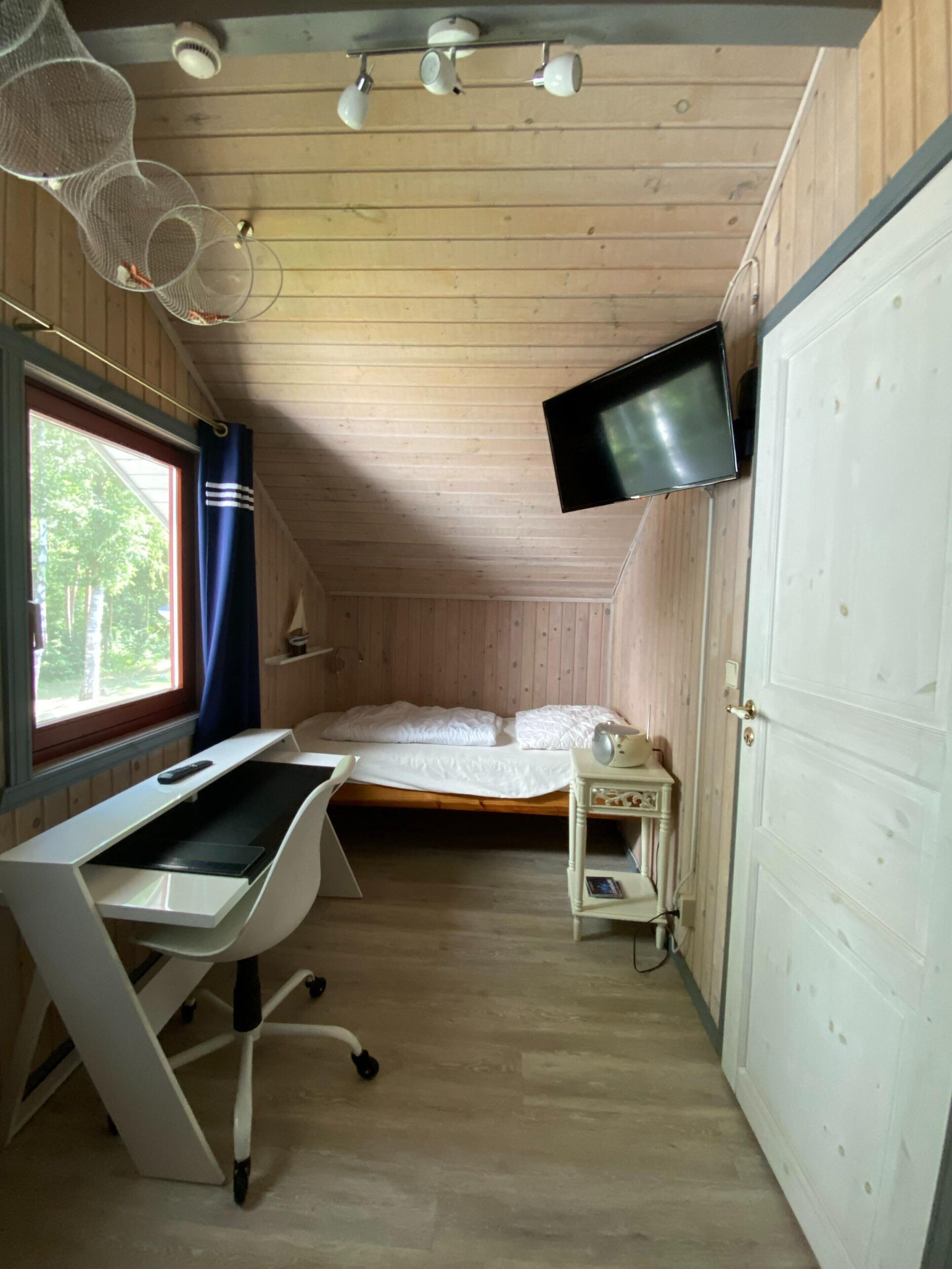 Ein kleiner, gemütlicher Raum mit einem Bett, einem Schreibtisch und einem an der Wand befestigten Fernseher.