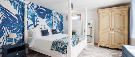 Un lit à baldaquin agrémenté d’un mur décoré de motifs de feuilles tropicales, une armoire en bois et un parquet à chevrons.