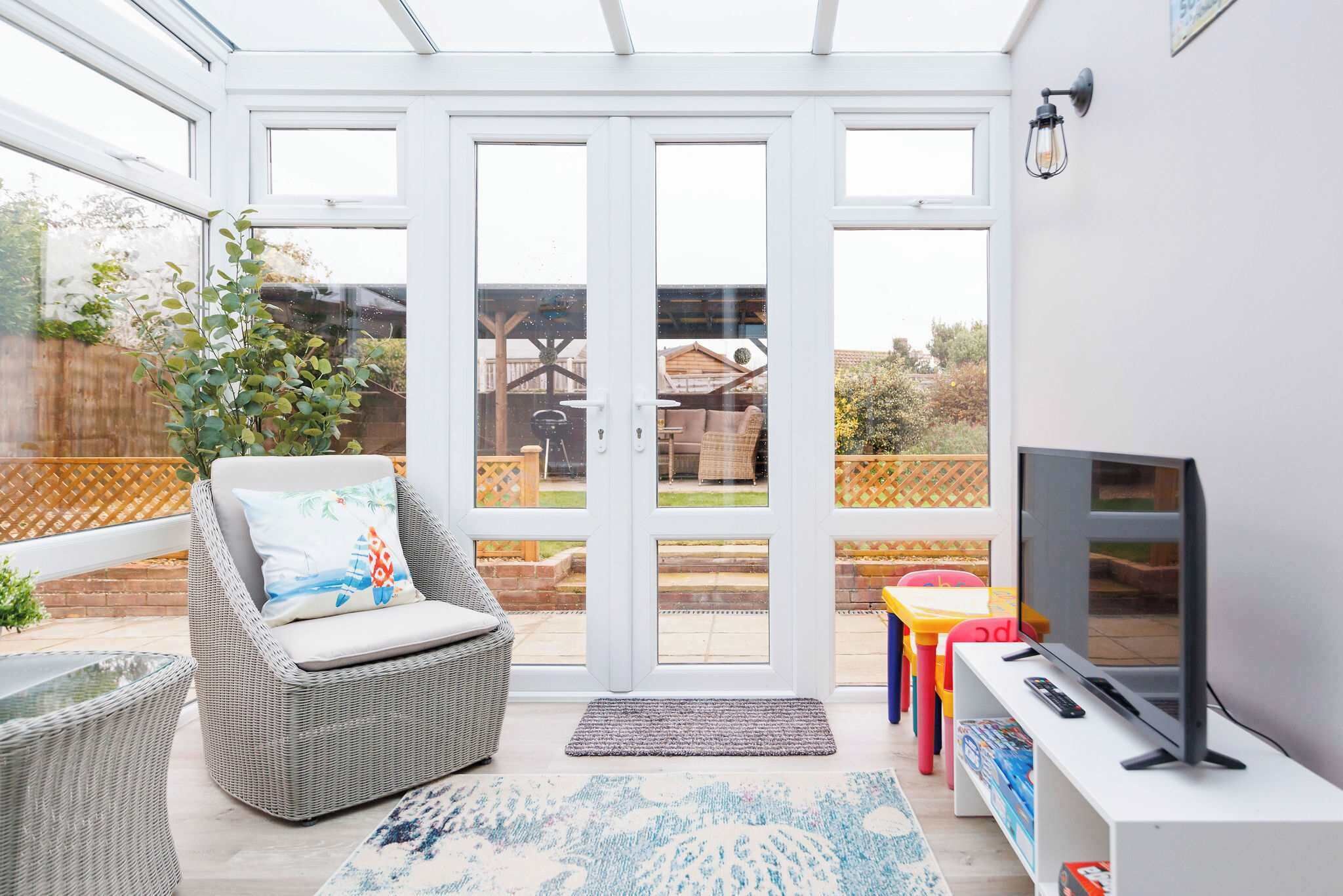 A conservatory with a glass door, wicker furniture, a television, and a small table with colorful chairs.