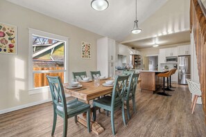 A dining area with a wooden table and chairs, a kitchen in the background, and a window with a view of a house.