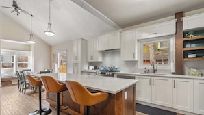 A modern kitchen with white cabinets, a large island with bar stools, a stove, and a window overlooking a garden. - Dino's Last Post | Sleeps 8 | Private Backyard! | (Drumheller)