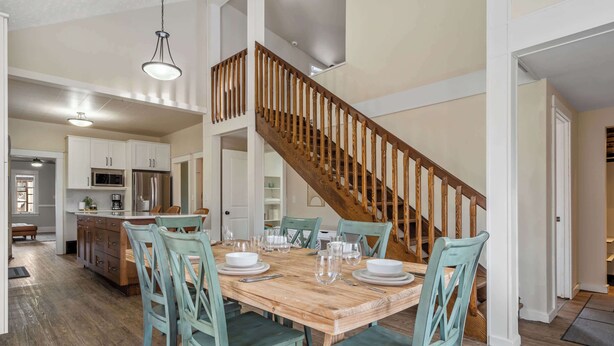 A dining area with a wooden table and chairs, a staircase, and a kitchen in the background. - Dino's Last Post | Sleeps 8 | Private Backyard! | (Drumheller)