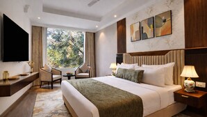 A hotel room with a bed, bedside tables, a TV, a desk, a chair, and a window with a view of trees.
