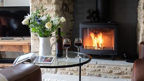 Ein gemütliches Wohnzimmer mit Kamin, einem Glastisch mit einer Zeitschrift, zwei Weingläsern und einem Blumenstrauß.