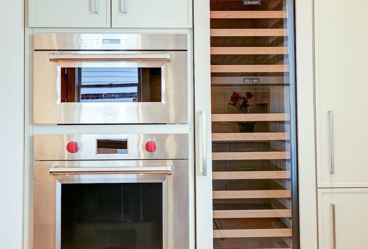 A kitchen with two stainless steel ovens, a wooden cabinet, and a glass-fronted shelving unit.