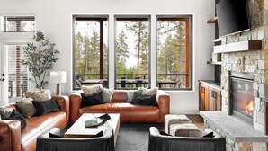 A cozy living room with a leather sofa, a fireplace, and large windows overlooking a forest.